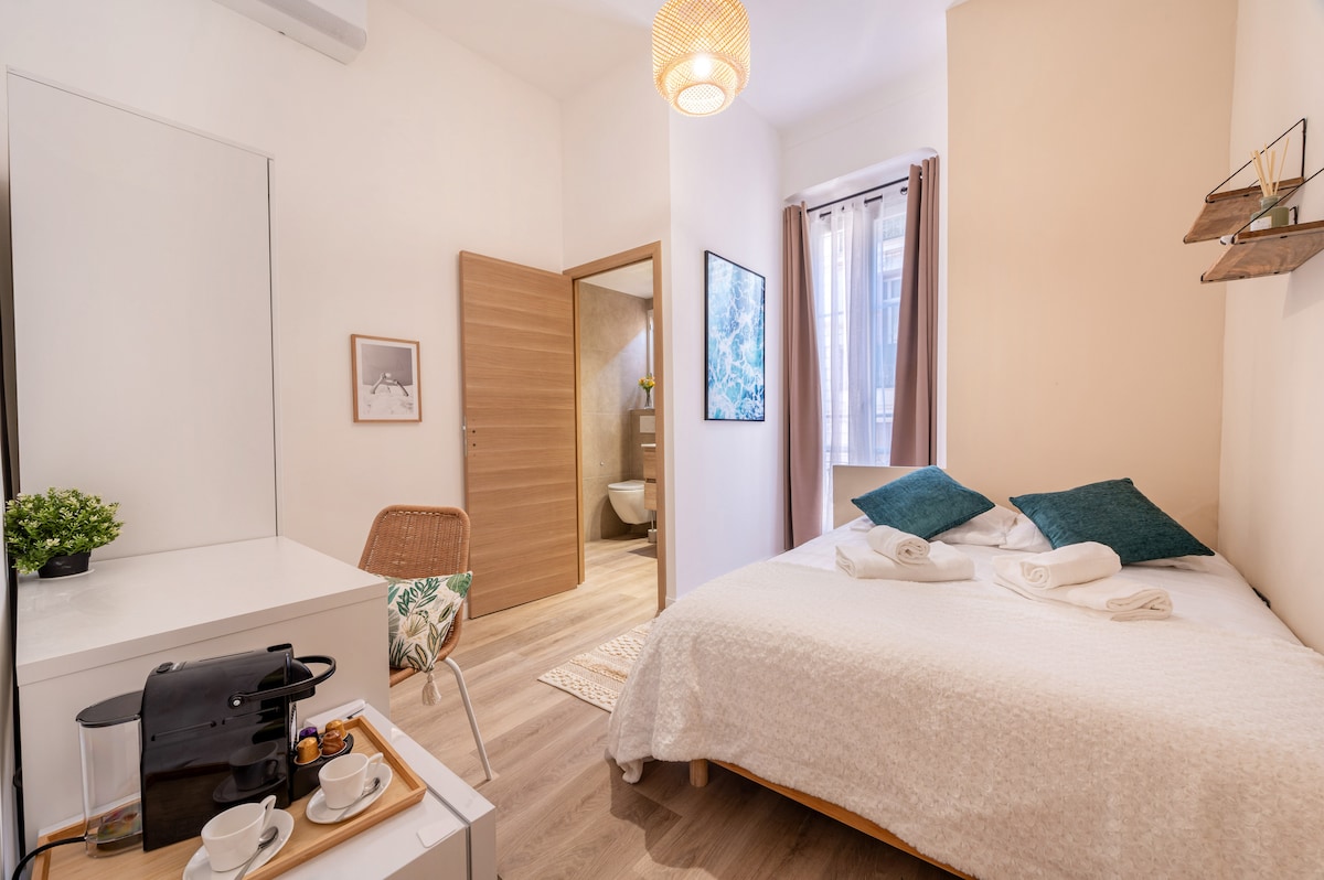 A well-lit room features a queen-size bed with white linens and decorative pillows. A desk with two cups and a coffee maker is present, alongside a chair. The room includes a glimpse of a private bathroom through the open door, and light curtains frame the window.