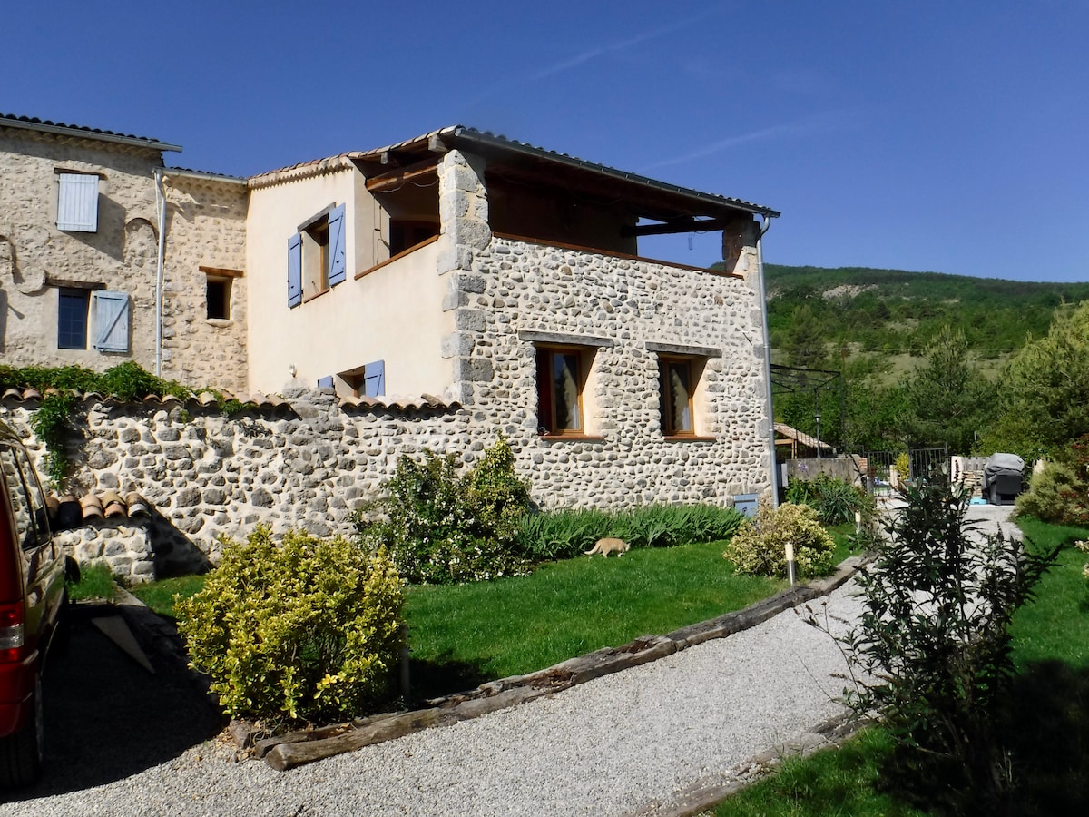 The rustic exterior of the barn is highlighted by natural stone walls and wooden shutters. A covered porch extends from the upper floor, offering a view of the surrounding landscape. Lush greenery and a gravel pathway lead up to the entrance, creating an inviting atmosphere.
