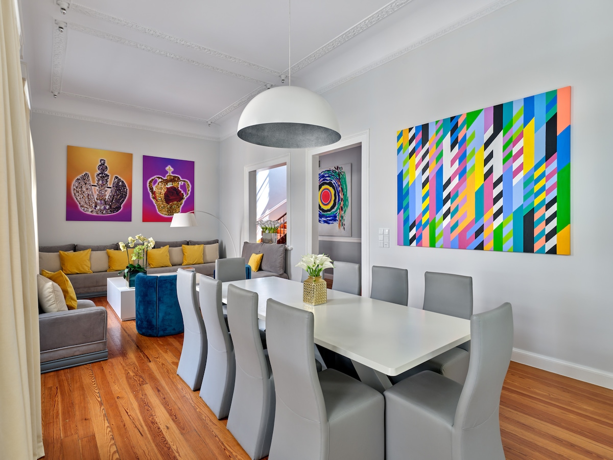 A spacious dining area showcases a modern white table surrounded by grey chairs. A comfortable sitting area with colorful cushions and large artwork can be seen in the background. Natural light fills the room, highlighting the elegant decor and wooden flooring.