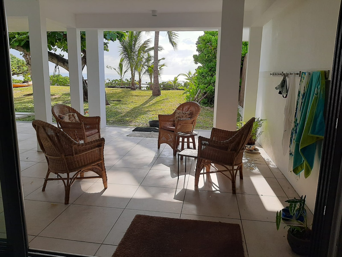 A shaded outdoor patio features four wicker chairs positioned around a central table, inviting relaxation. Lush greenery and palm trees are visible in the background, enhancing the tranquil setting. The tiled floor provides a cool surface beneath the seating area, ideal for enjoying the warm climate.