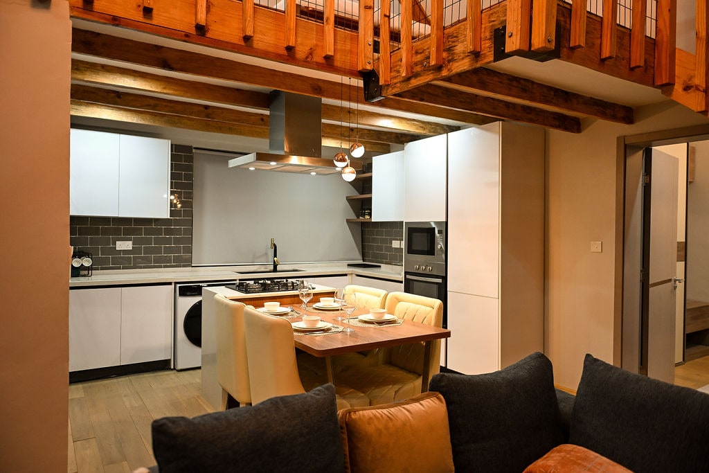 An open-plan kitchen is showcased with modern appliances and sleek cabinetry. A dining table is set with tableware, while soft lighting from pendant fixtures adds warmth. Wooden beams highlight the ceiling, complementing the cozy seating area with a mix of textured cushions.