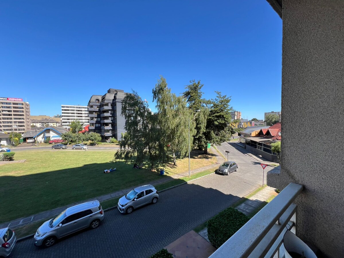 A view from the balcony shows a grassy area with trees and various vehicles parked along the street. The clear blue sky complements the scene, while nearby buildings form part of the urban landscape.