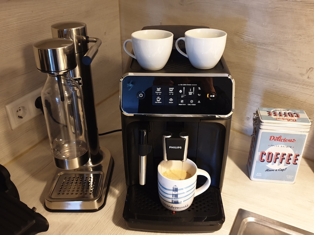 An automatic coffee machine is displayed alongside a glass coffee press. Two white coffee cups sit atop the machine, while a striped mug is positioned below the spout, filled with freshly brewed coffee. A tin of coffee labeled 'Delicious' is visible in the background.