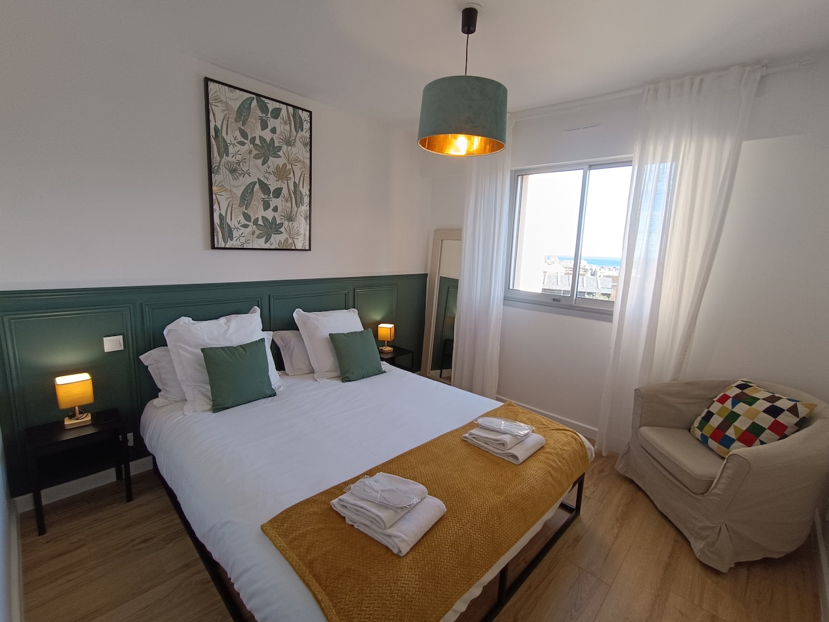 A well-lit bedroom features a spacious bed with two green accent pillows and crisp white linens. A mustard-colored throw blanket is neatly draped across the bed. A cozy armchair sits in the corner, complemented by a large window allowing natural light, with light curtains fluttering gently.