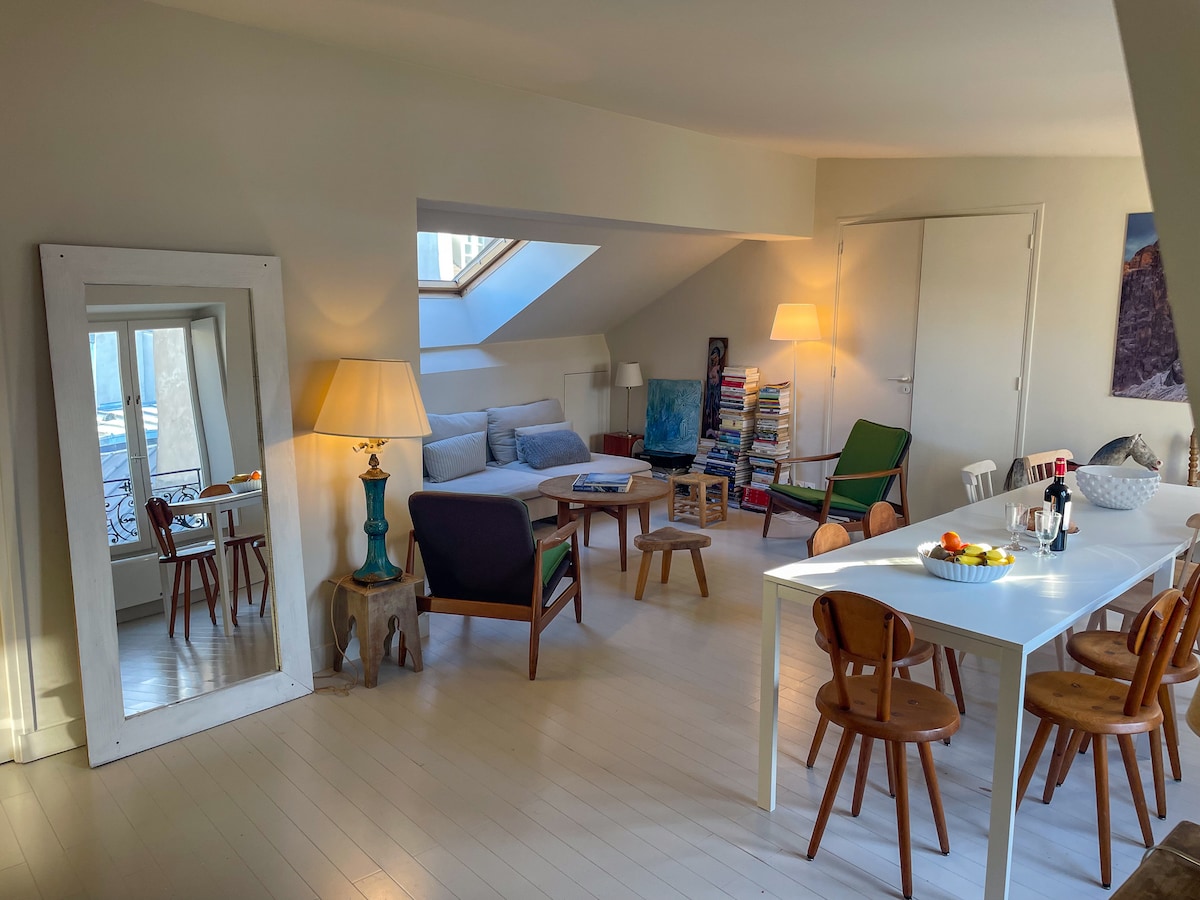 A bright living area features large windows with natural light flooding in. A spacious sofa is complemented by a reading chair and a small coffee table. A dining table with chairs is set for a meal, while a decorative mirror reflects the room's inviting atmosphere.