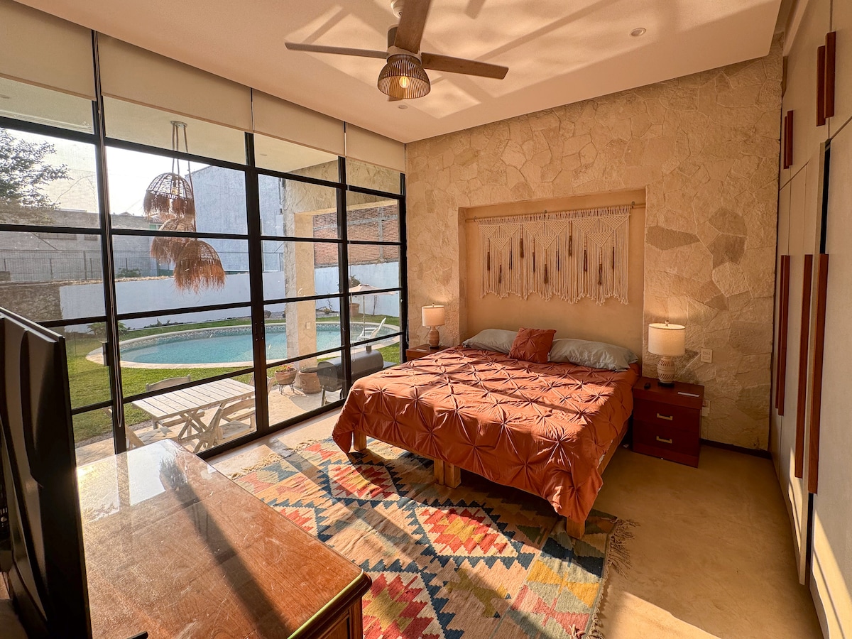 A spacious bedroom features a king size bed with a textured orange coverlet. Large windows allow natural light to illuminate the room, offering views of the private garden and pool. A wooden dresser and bedside tables add functionality, while a fan provides added comfort.