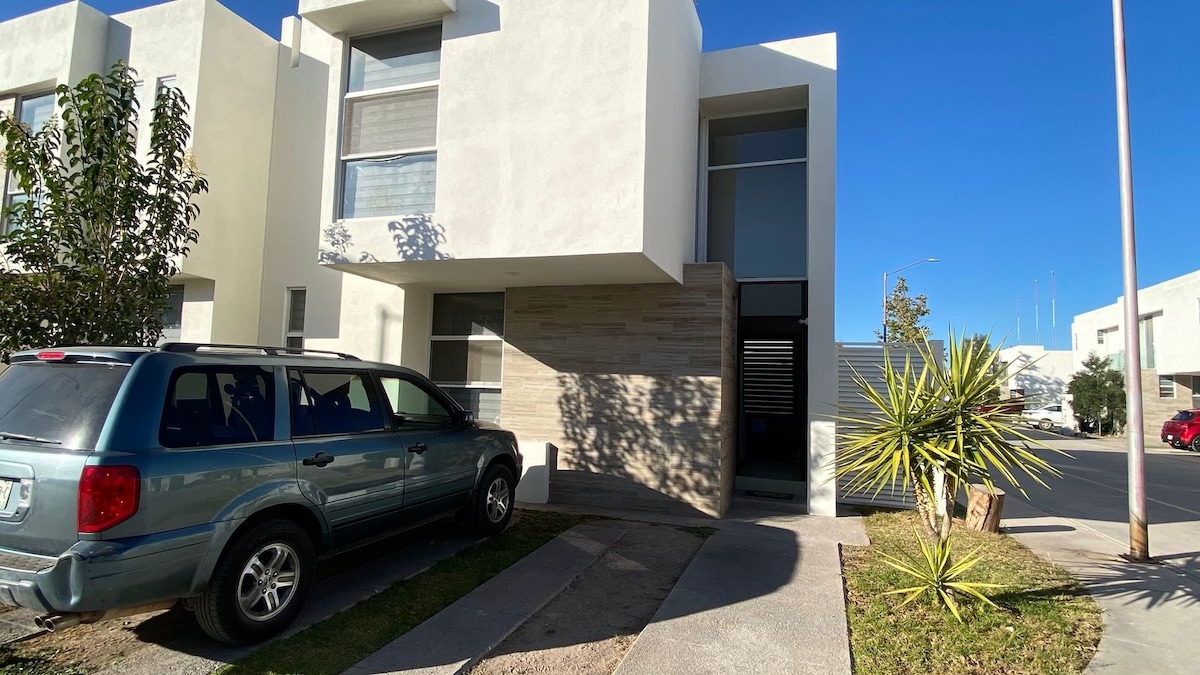 A modern building is showcased with minimalistic architectural details. A compact vehicle is parked on the driveway, accompanied by native plants and neatly maintained landscaping. Bright natural light illuminates the exterior, enhancing the crisp lines and contemporary design.