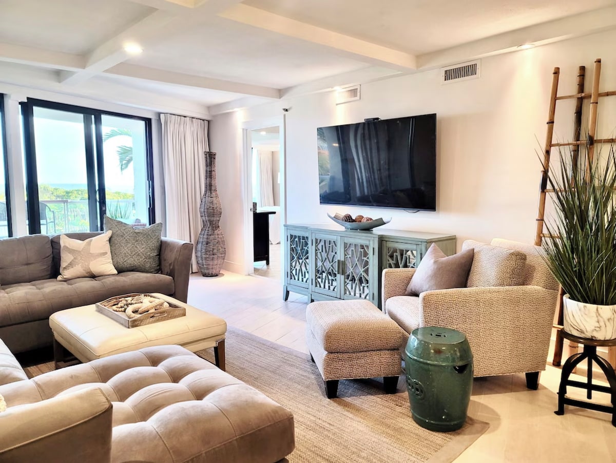 A bright and modern living area is presented, featuring a spacious seating arrangement with a light-colored sectional sofa and a cozy armchair. A large television is mounted on the wall, while a woven basket and decorative items adorn the console table. Natural light fills the room through sliding glass doors.