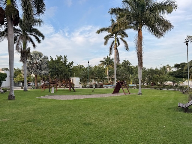 A well-maintained green space features manicured lawns, palm trees, and playground equipment. Benches are positioned along the pathways, providing seating in a peaceful outdoor environment suitable for families and friends. The setting is well-lit, enhancing safety and accessibility.