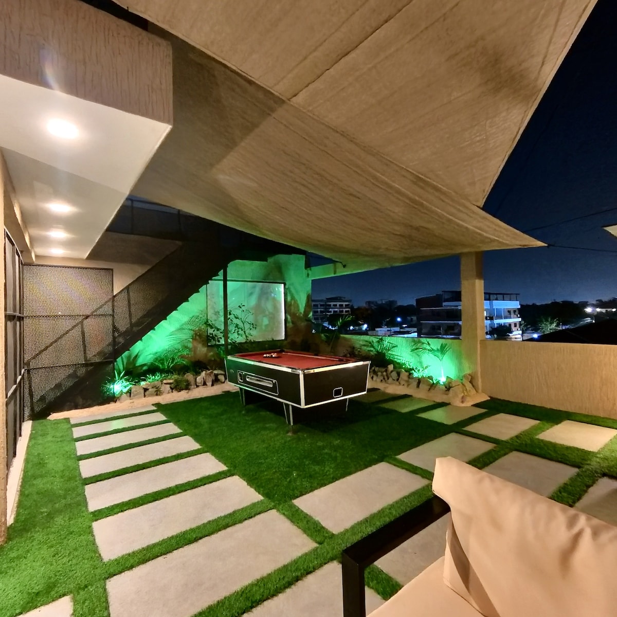 An outdoor lounge area showcases a pool table set against a backdrop of well-maintained greenery and soft illumination. The space is neatly arranged with stone pathways and reflected light from below, providing a serene atmosphere for relaxation or entertainment.