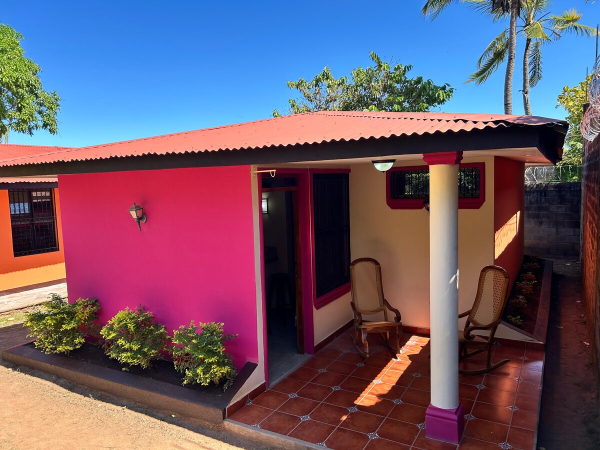 The exterior of the house features a colorful facade with pink and orange accents. Two wicker chairs are positioned on a tiled porch, surrounded by small greenery. A clear sky and palm trees create a bright, inviting atmosphere.