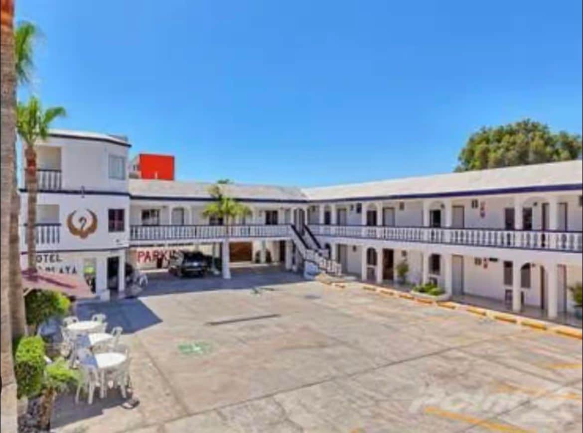 A central courtyard is surrounded by a two-story building, with staircases leading to the upper level. Potted plants add greenery, while a few outdoor tables and chairs are positioned on one side. Bright blue sky creates a vibrant backdrop for the scene.