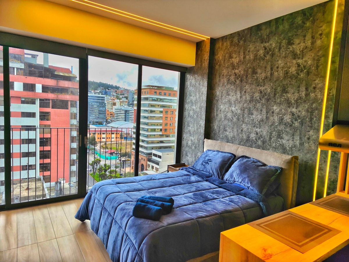 A modern bedroom features a spacious bed dressed in rich blue linens, complemented by a neatly placed towel. Floor-to-ceiling windows invite natural light, offering an expansive view of the cityscape beyond.