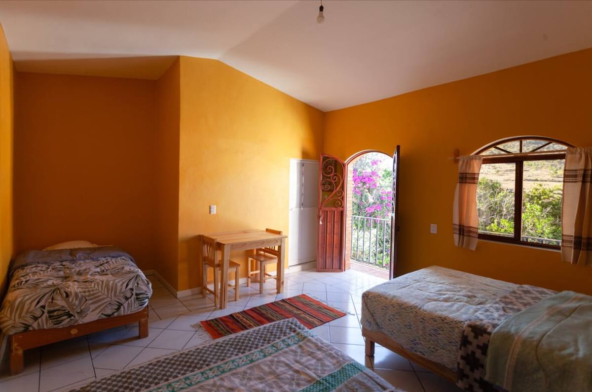 A spacious room features warm yellow walls and a mix of bedding options, including a single bed and two larger beds. A small dining table with chairs is positioned next to an open door, providing a view of a vibrant outdoor garden.