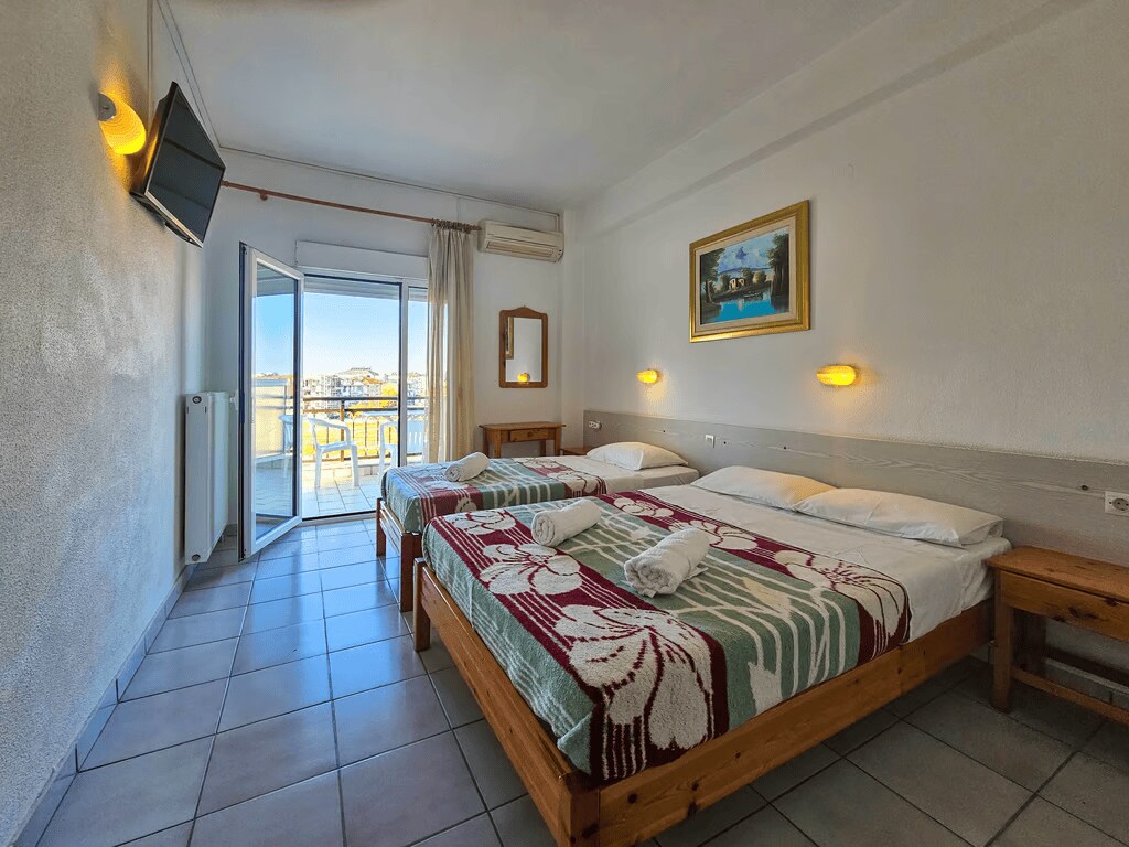 A light-filled triple room is presented with two beds, one double and one single, each adorned with fresh linens. A flat-screen TV is mounted on the wall, while large sliding doors open to reveal a street view, enhancing the spacious atmosphere.