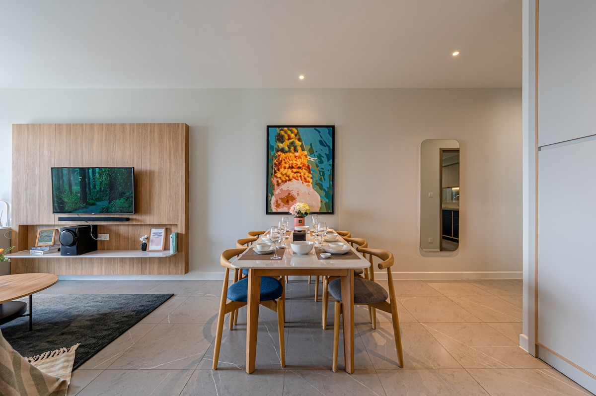 The dining area features a large stone table set for six, complemented by modern chairs in various colors. A decorative painting hangs on the wall, and an LED TV is mounted on a wooden unit, enhancing the space’s contemporary design.