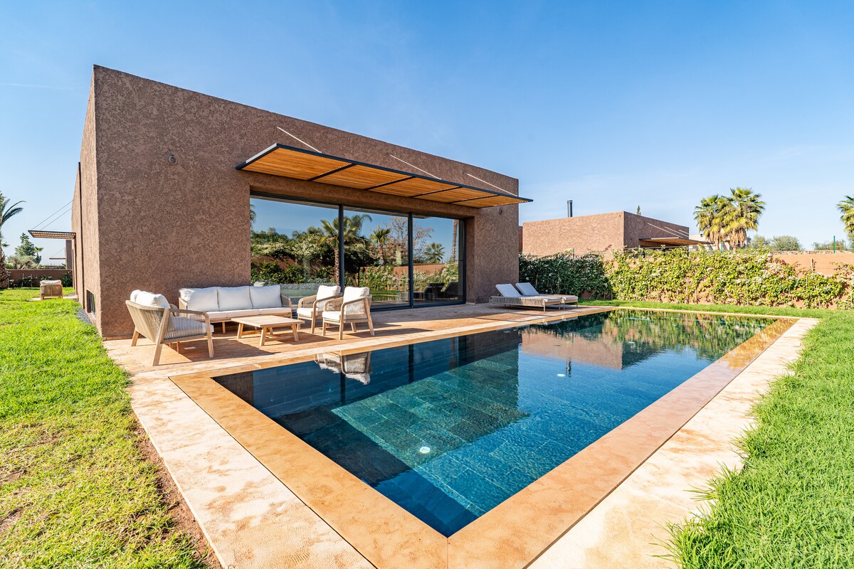 A modern villa features a spacious outdoor area with a private swimming pool surrounded by lush green grass. Comfortable seating is arranged next to the pool, with large windows providing a view of the serene landscape. Clear blue skies enhance the tranquil ambiance.
