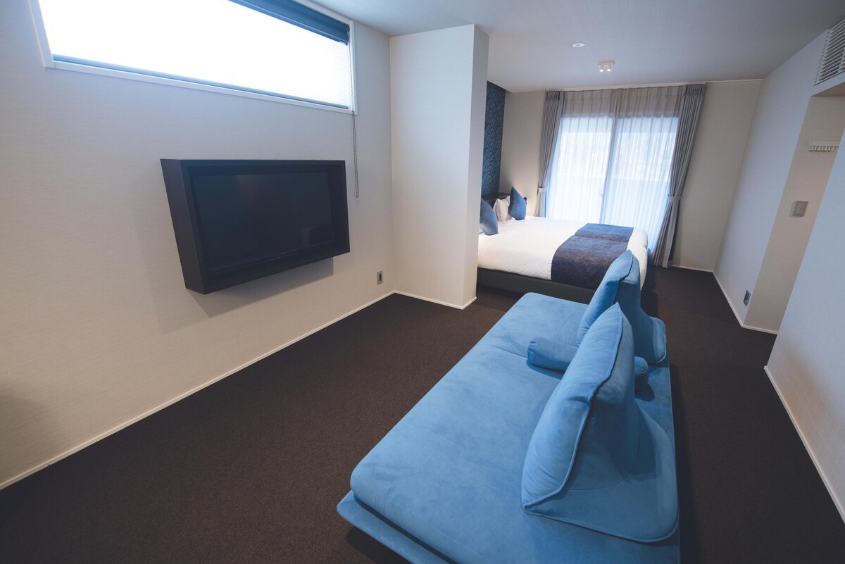 A spacious room features a cozy bed with a dark bedspread and crisp white linens. A blue couch is positioned near a television mounted on the wall. Natural light filters through the sheer curtains, highlighting the neutral tones of the room.