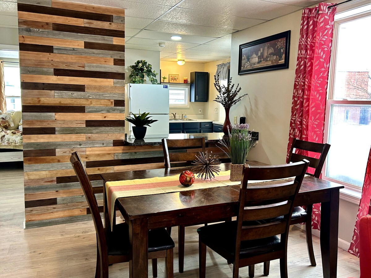 A dining area is shown with a wooden table surrounded by four chairs, complemented by a decorative centerpiece of flowers. A textured wood accent wall serves as a backdrop, while a brightly colored window treatment adds a pop of color. A small kitchen area is partially visible.