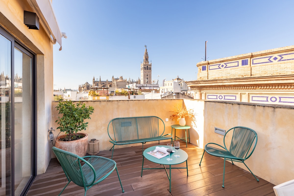 A private terrace is presented with green metal furniture, including seating and a table. The views feature the skyline of Seville, highlighted by the Cathedral's tower. A potted plant adds a touch of greenery, complementing the warm tones of the surrounding walls.