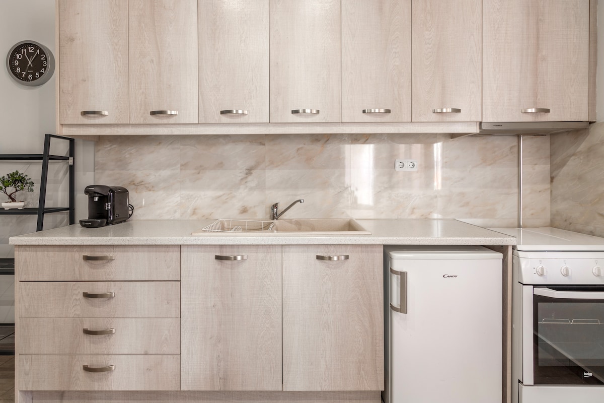 A modern kitchen features light-toned cabinetry and a smooth countertop. Essential appliances, including a refrigerator, stove, and coffee maker, are neatly arranged. A subtle marble backsplash adds a touch of elegance, while ample storage space is provided with multiple drawers and cabinets.
