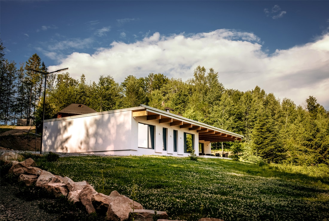A modern structure is positioned within a lush green landscape, featuring a long overhang that provides shade. The building's facade is white with multiple doors leading to the outdoors, surrounded by a natural setting of trees and a grassy area.