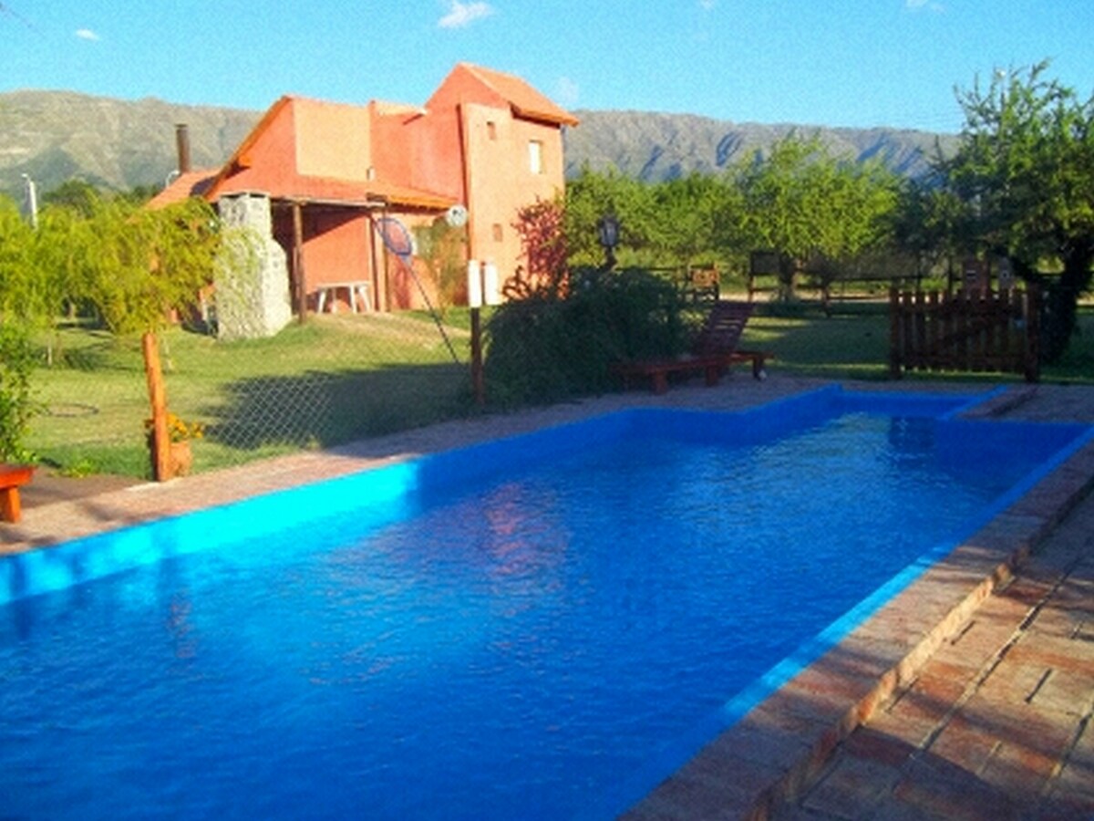 A clear blue swimming pool is situated in front of a rustic red building surrounded by lush greenery and mountains in the background. Lounge chairs are placed nearby, inviting relaxation in a tranquil outdoor setting.