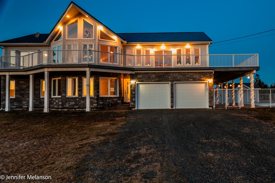 The exterior of the home is illuminated at dusk, highlighting the stone facade and large windows. A spacious deck extends above, providing elevated views. Two garage doors are visible, and the surrounding grounds feature a gravel driveway and outdoor lighting.