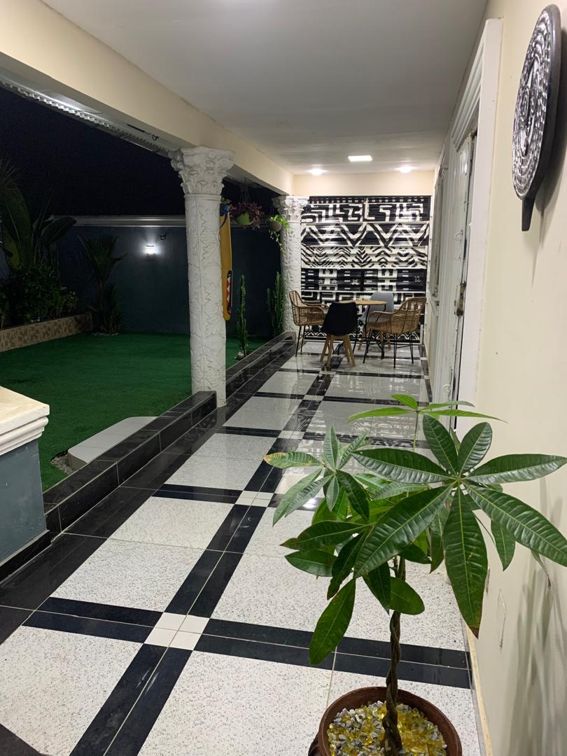 A well-maintained corridor is framed by marble columns, featuring a green grass area and a modern outdoor dining setting. Decorative plants enhance the space, while a geometric patterned wall adds a contemporary touch. Soft lighting creates a calming ambiance.