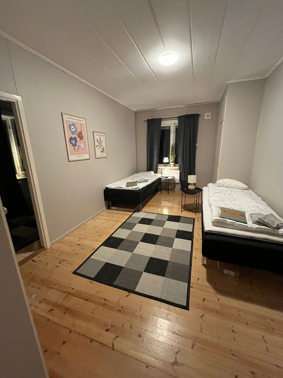 A bright bedroom features two single beds arranged with neatly made linens. A small table is positioned between the beds, and a patterned area rug covers the polished wooden floor. Natural light is provided by a window showcasing simple decor on the walls.
