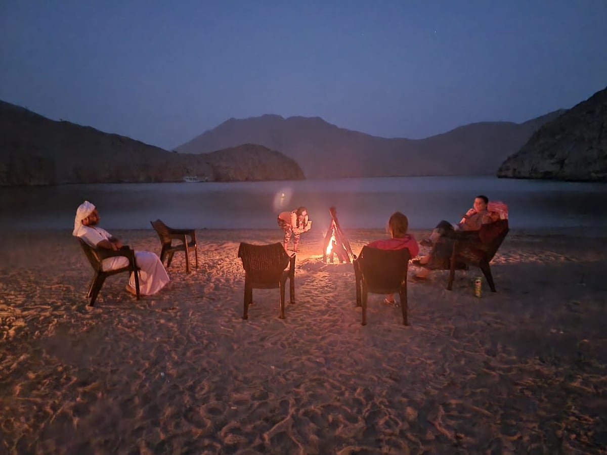 Several chairs are arranged around a glowing campfire on a sandy beach, with mountains visible in the background. The evening sky is darkening, and the calm water reflects faint light from the fire, creating a serene atmosphere.