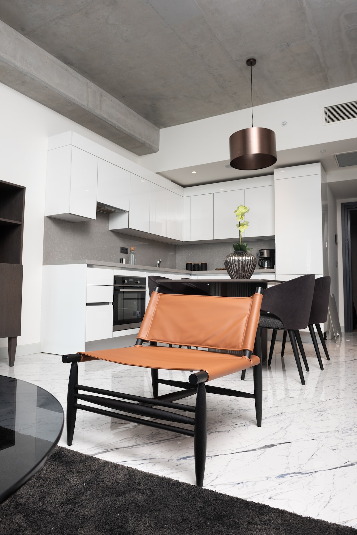 A modern kitchen space features sleek cabinetry and stainless steel appliances. A stylish orange chair is placed in the foreground, while a dining area with dark chairs is visible. Natural light illuminates the marble flooring, enhancing the overall contemporary design.