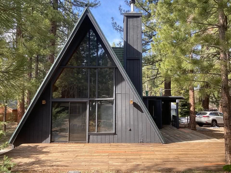 A modern A-frame house is surrounded by tall pine trees, showcasing a wooden deck that extends from the front. Large glass panels dominate the front facade, allowing natural light to fill the interior. The structure features a chimney and a dark exterior finish.