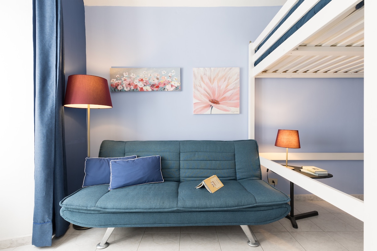 A comfortable seating area features a blue sofa bed adorned with cushions. A book lies open on the couch, and is complemented by decorative wall art above. Warm lighting is provided by two lamps, enhancing the inviting blue and white color scheme.
