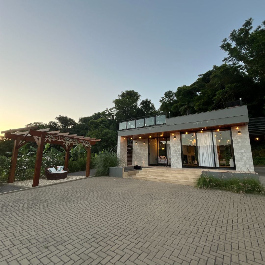 A modern cabin is set amidst lush greenery, featuring a stone and wood exterior. A covered terrace is shown, alongside a pathway leading to large glass doors that open into the interior. Soft lighting enhances the inviting ambiance as the sky transitions to twilight.