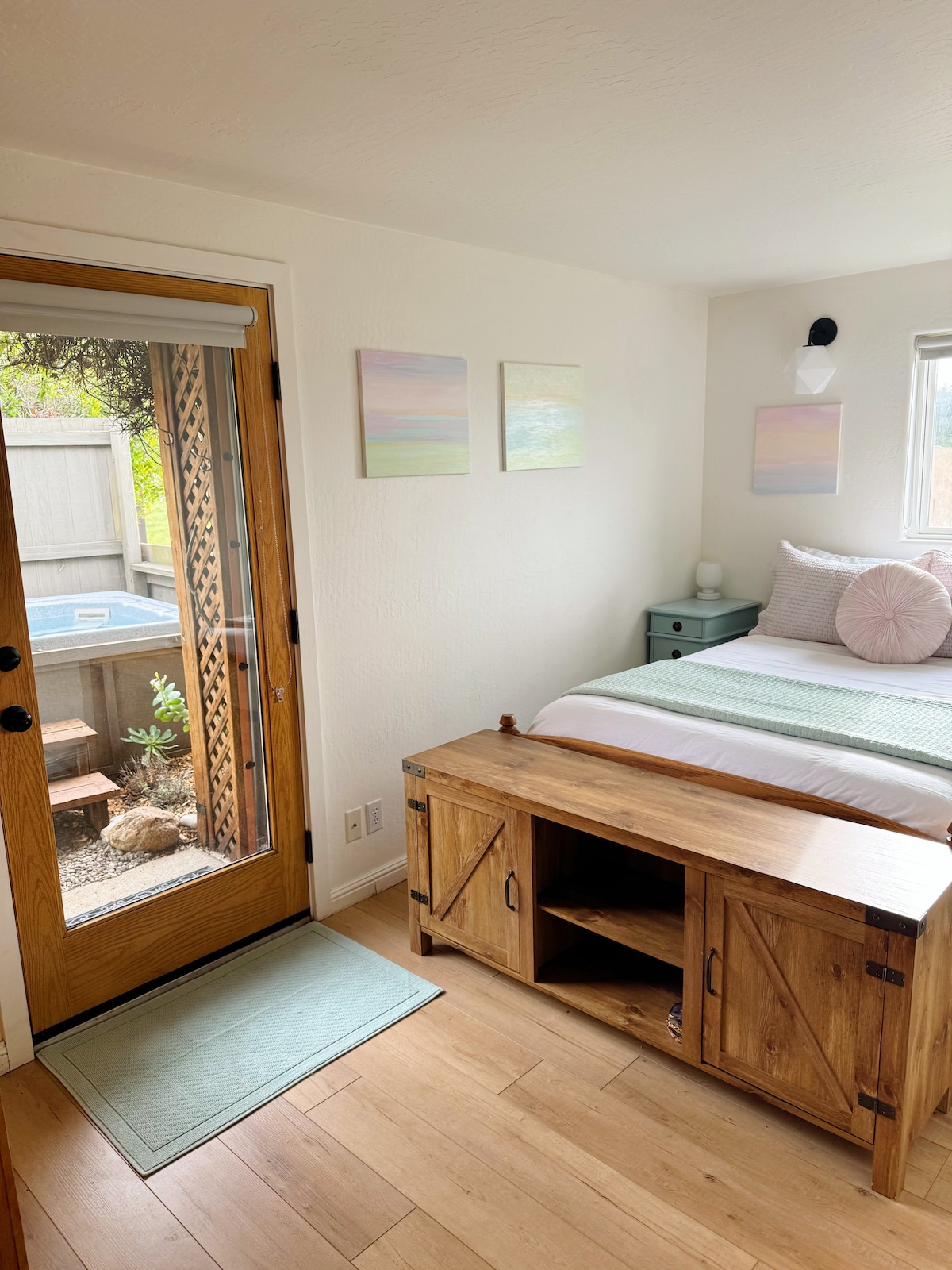 A bright and airy room features a spacious bed with light bedding, complemented by a wooden bench at the foot. A door leads to an outdoor space, framed by light walls and adorned with landscape art. Natural light enters through the nearby window.