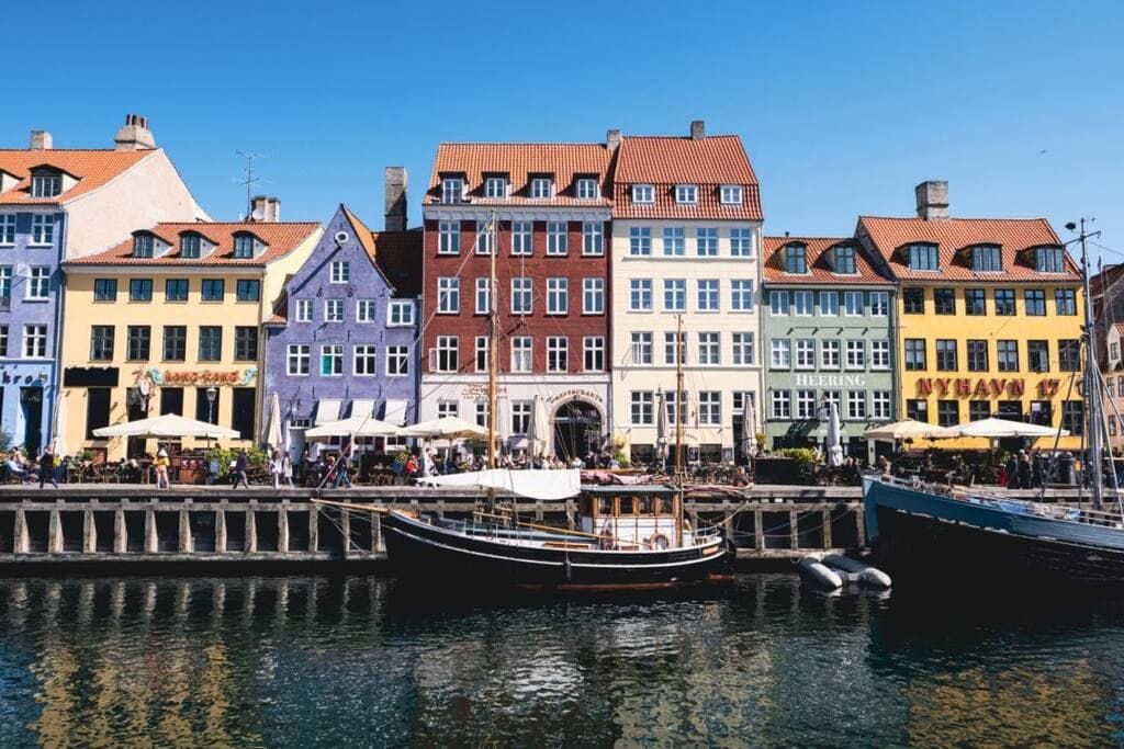 A row of colorful historic buildings faces a calm waterfront. The structures feature various vibrant hues, including purple, red, and yellow, with distinct roofs. Outdoor seating areas with umbrellas are visible, offering guests a scenic view of the canal and surrounding architecture.