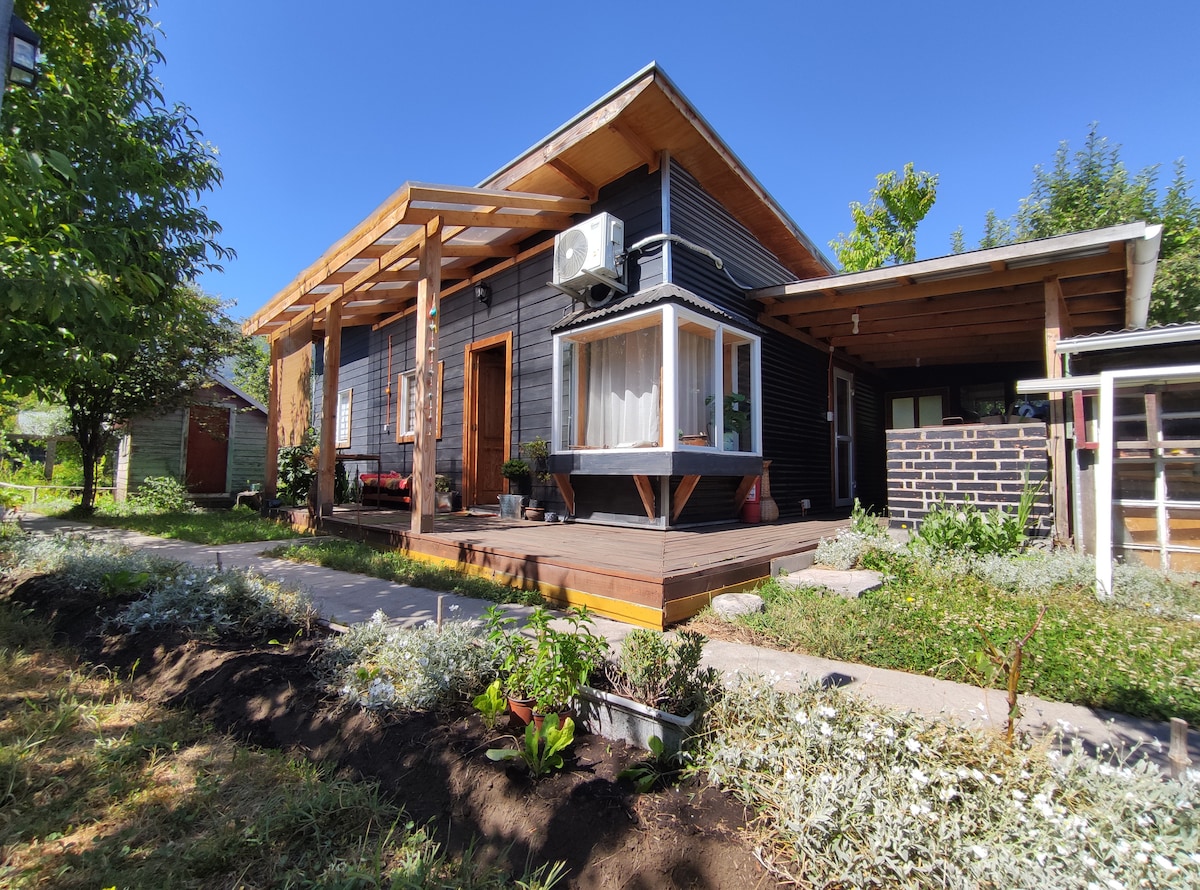 A modern cabin exterior features a front porch with a wooden frame, providing shade. Large windows invite natural light, while a mounted air conditioning unit is visible. The landscaped yard includes various plants and flowers, set against a clear blue sky.