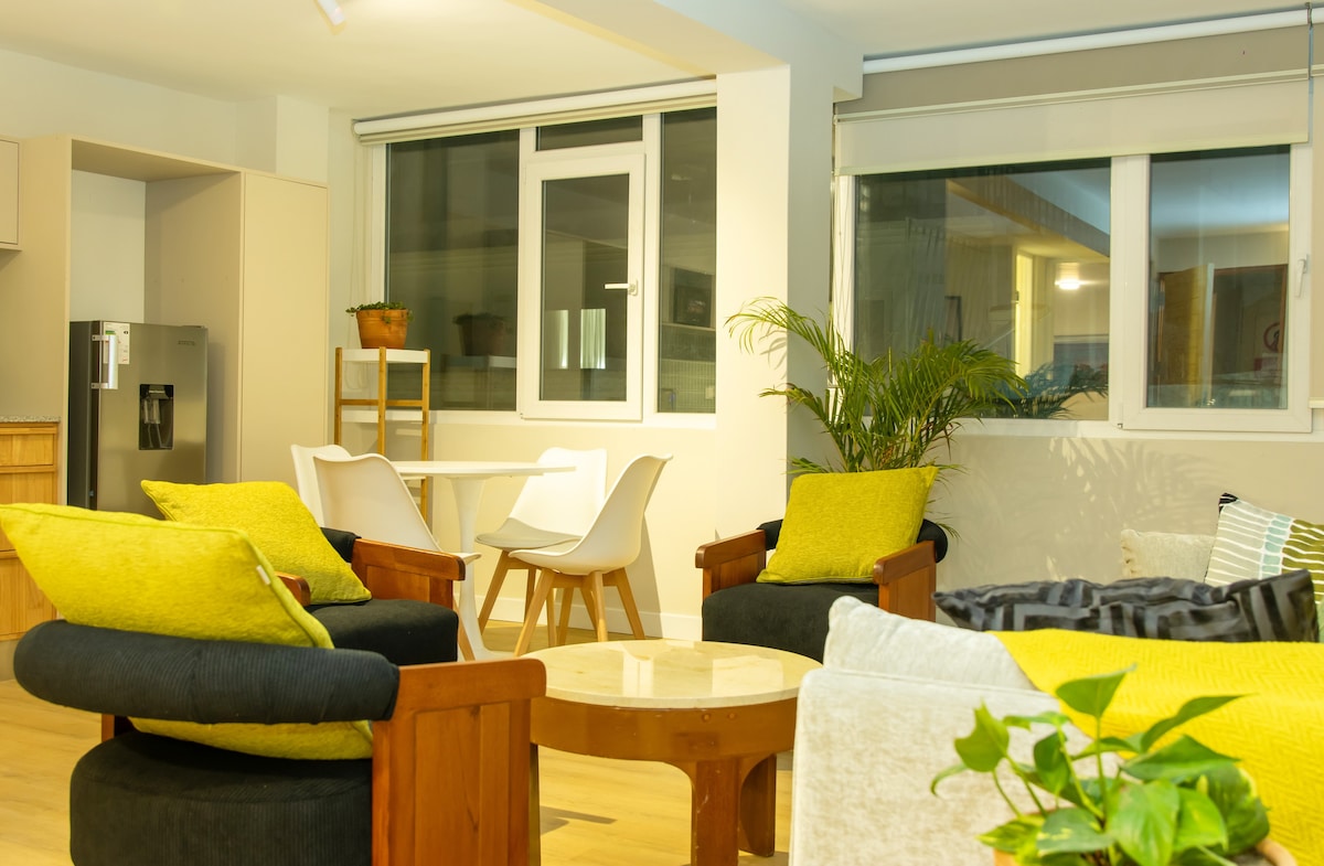 A spacious living area is featured, showcasing a circular wooden table surrounded by white chairs. Comfortable armchairs with vibrant yellow cushions are positioned nearby. The room is well-lit, with large windows allowing natural light and a view of greenery from potted plants.