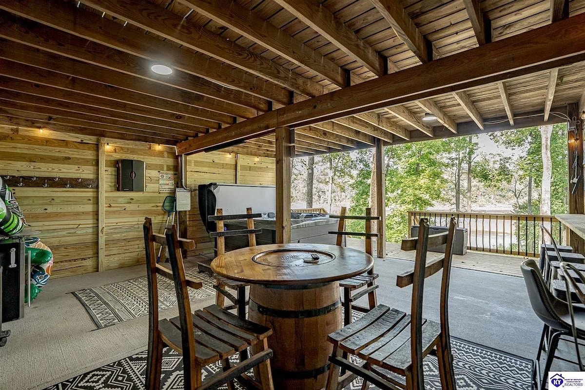 An outdoor dining area is showcased under a wooden roof, featuring a round table crafted from a barrel surrounded by sturdy wooden chairs. A grill is visible in the background, with trees providing a serene backdrop. Natural light filters in, enhancing the space's openness.