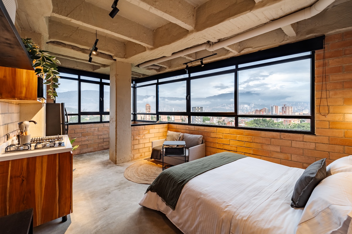 An open-plan bedroom features a spacious bed, complemented by large windows that offer expansive city views. Natural light fills the room, while minimalistic decor and a cozy sitting area are accentuated by warm tones and an inviting rug.