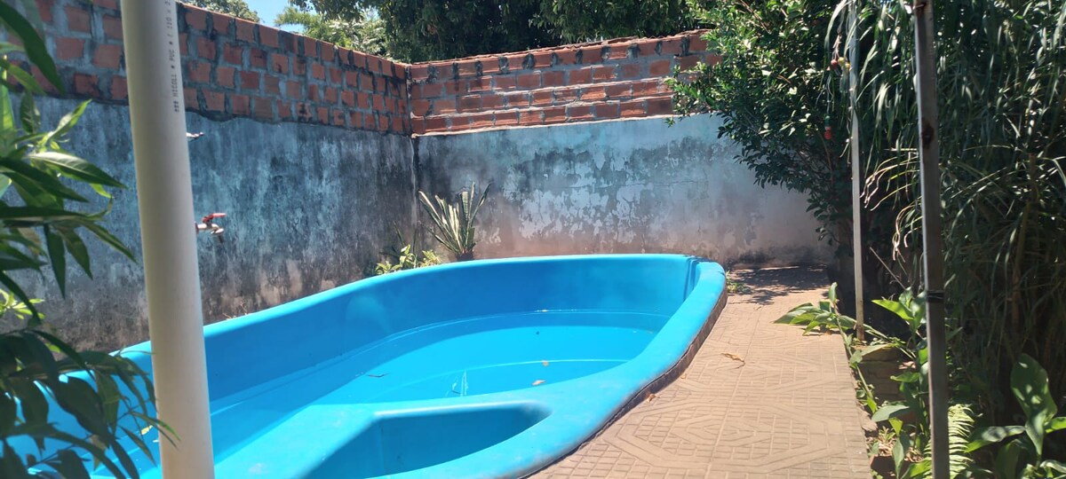 A turquoise swimming pool is surrounded by a clean, paved area, complemented by green plants. The pool's shape is distinct, and the backdrop features a textured brick wall alongside a partially painted surface, creating a serene outdoor space.