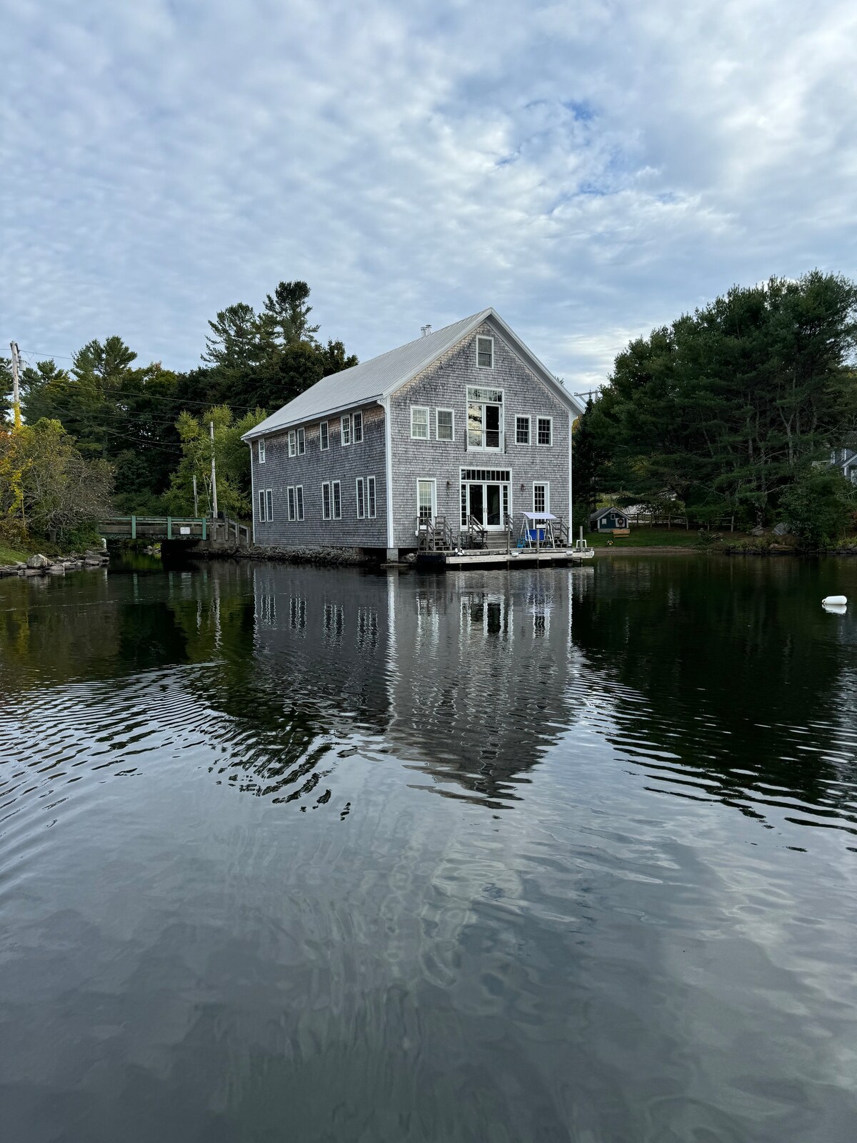 A charming shingle-clad home is depicted standing on the edge of Damariscotta Lake. The calm water reflects the structure and surrounding trees, creating a serene atmosphere. Soft clouds are visible in the sky, adding texture to the scene.