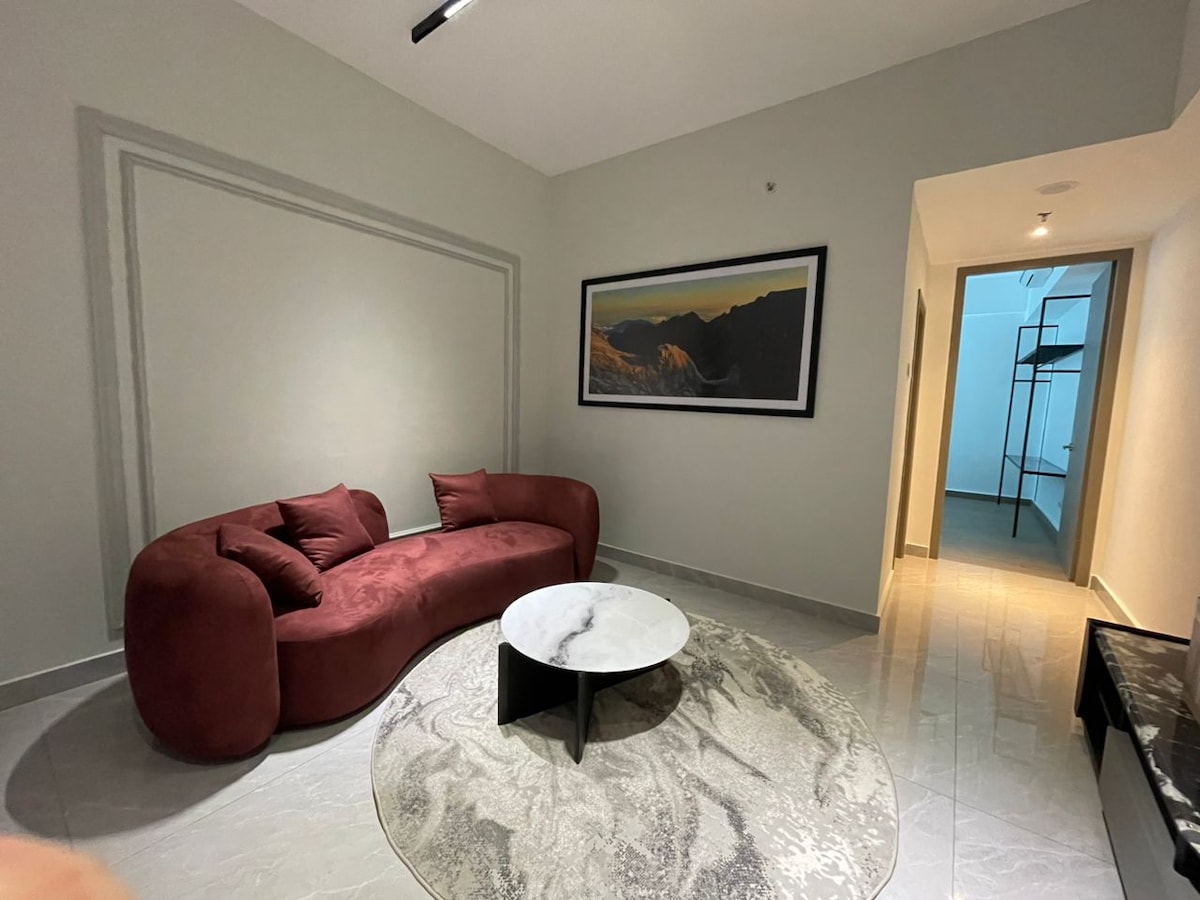 A modern living area features a deep red, curved sofa paired with a round coffee table. A soft area rug grounds the space, which is illuminated by natural light. A large artwork depicting a mountain landscape adorns the wall, adding depth to the room.
