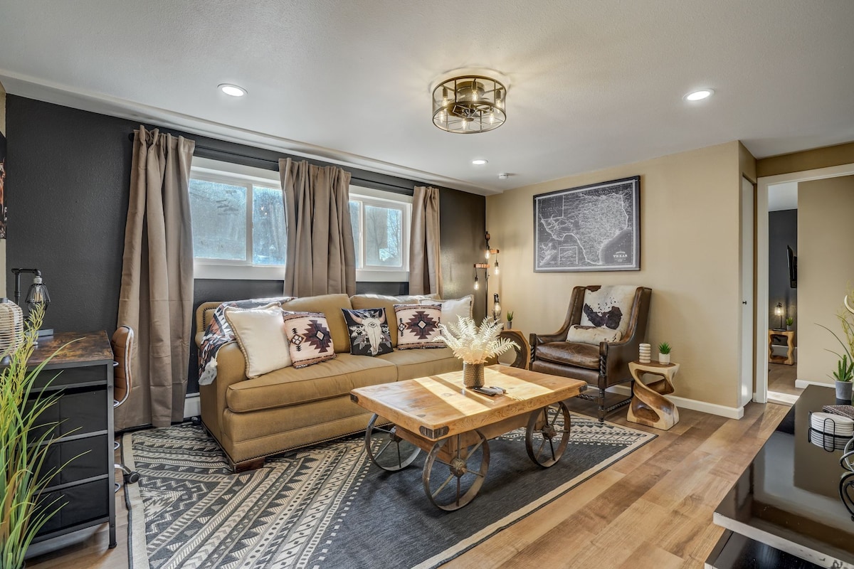A cozy living area features a beige sofa adorned with decorative cushions, complemented by a rustic coffee table with wheels. Natural light enters through large windows, which are framed by chic curtains. A stylish armchair adds to the inviting atmosphere, while wall art enhances the decor.