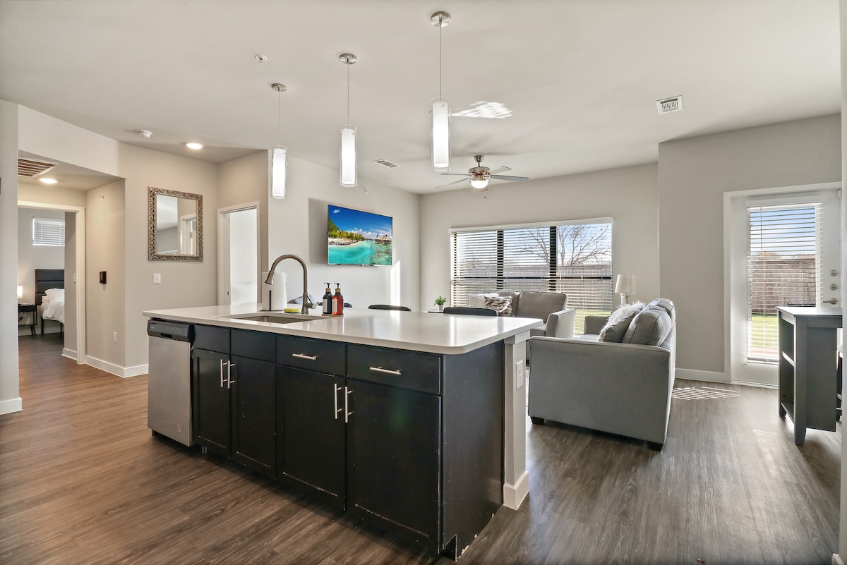 An open living and dining area is showcased with a large kitchen island featuring modern fixtures. Natural light floods through multiple windows, illuminating the cozy seating arrangement and a mounted television. A door leads to a patio area outside, enhancing the spacious feel.