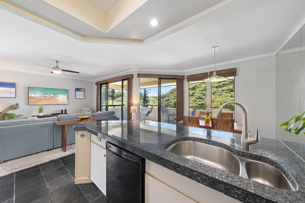 A contemporary kitchen with dark granite counter tops is visible in the foreground. Beyond the kitchen, a spacious living area features a couch and dining table, with large sliding glass doors offering views of a green landscape and outdoor seating on the lanai.