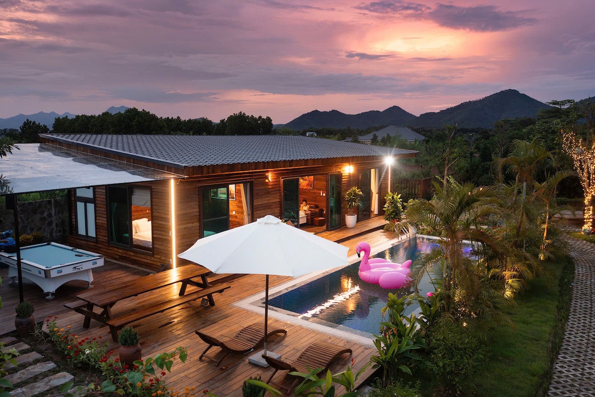 An inviting outdoor space is depicted, featuring a swimming pool with a pink flamingo float. Surrounding the pool are sun loungers and a large umbrella. The wooden deck is complemented by tropical plants and mountains in the background, all under a vibrant sunset sky.