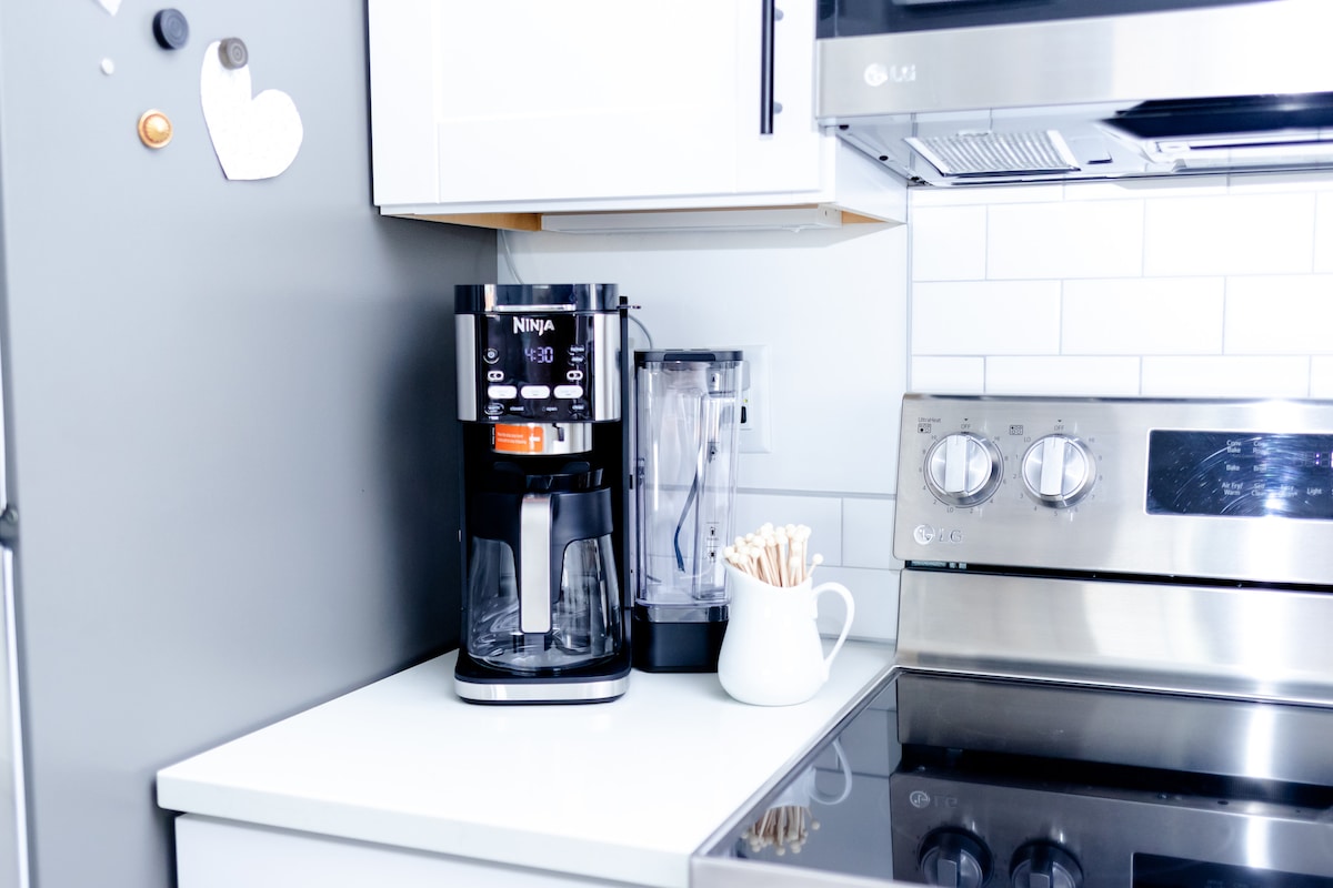 A modern kitchen corner includes a sleek coffee maker and blender, complemented by a white pitcher holding wooden stirrers. A stainless steel oven and a clean countertop enhance the area’s bright and inviting look, with tile backsplash adding a touch of simplicity.