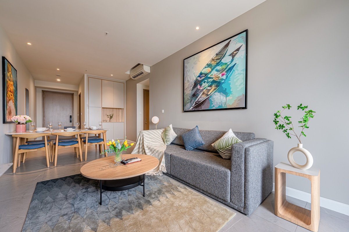 A spacious living area features a cozy sofa with decorative pillows and a round coffee table. A modern dining table is seen in the background, complemented by an artwork on the wall. Natural light streams through large windows, highlighting the contemporary decor.